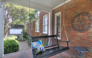 Red brick home porch with brick floor black hanging swing and black metal side table with shed and bird fountain in the background