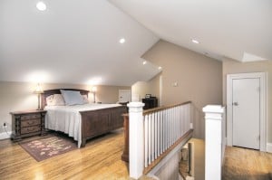 Bedroom with slanted ceiling and recessed lighting with light brown walls and wood floors white railing and wood banisters on stairs going down