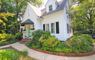 White siding two story home with black shutters and blue front door with black awning over it brick lined cement pathway