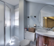 Bathroom with light blue walls white baseboards white tile floors white toilet brown cabinet with white sink and large round mirror above it