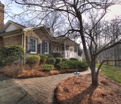 Brown brick home with black shutters front porch with white railing and columns brick path in front yard