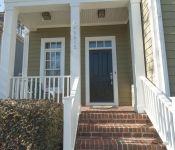 Front porch of home with green siding black front door red brick stairs to front porch white columns and white railings around porch