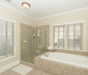 Bathroom with beige walls white baseboards two windows with built in white and brown blinds brown tile flooring and tile in shower and around bathtub