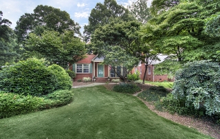 Brick home with green shutters and green front door large trees and bushes around it