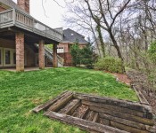Back of a brown brick two story home with green grass and trees on the border of the yard back patio and second floor gray painted deck