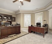 Home office with light brown walls white molding light carpet windows behind desk two wood desks large wood bookshelf