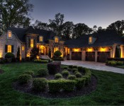 Front of large three story brick and stone home with three car garage and manicured front lawn at night with lights shining on home