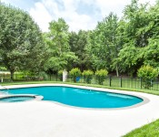 Backyard with round built in pool and hot tub cement around it fenced in with black iron fence green grass and trees behind fence