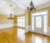 Empty dining room with wood floors yellow walls white chair molding three windows with white blinds chandelier in the middle of the room open to front entry way