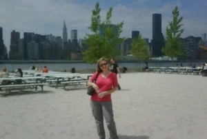Beach with skyline behind it picnic tables and woman wearing a pink top and gray pants smiling for the camera