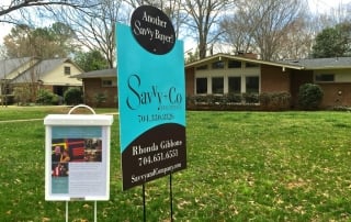 Brown brick home with large green grass front yard and brown and blue Savvy and Co Real Estate sign in front of it