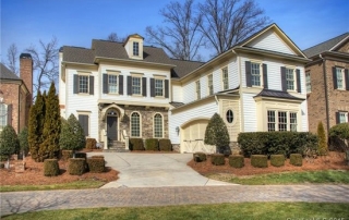 Large three story white siding home with black shutters cream and stone accents brick paved sidewalk in front manicured bushes and trees in front yard