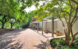 Large front porch of brown and white home with potted flowers tree lined brick pathway