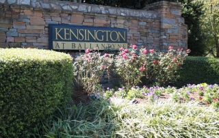 Stone wall with black and gold sign on it Kensington at Ballantyne on it with flowers and bushes planted in front