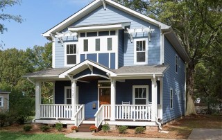 Two story blue siding home with white trim and front porch with white railing and columns brown wood front door driveway on the right large tree in backyard