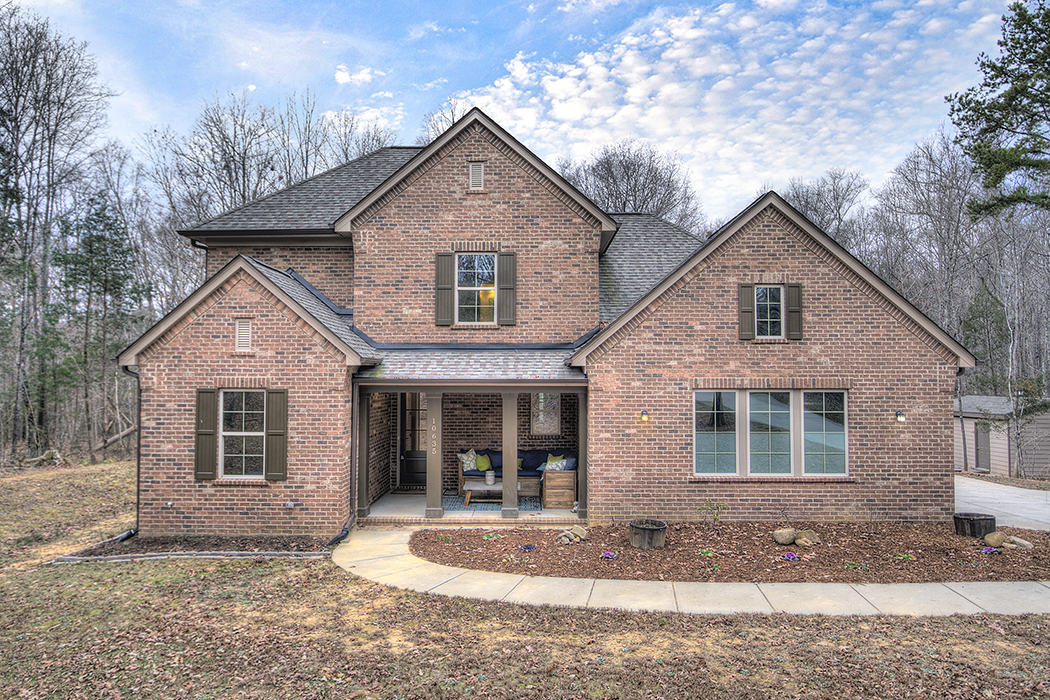 Two story brown brick home with brown shutters front porch with brown columns cement walkway to front door trees in backyard