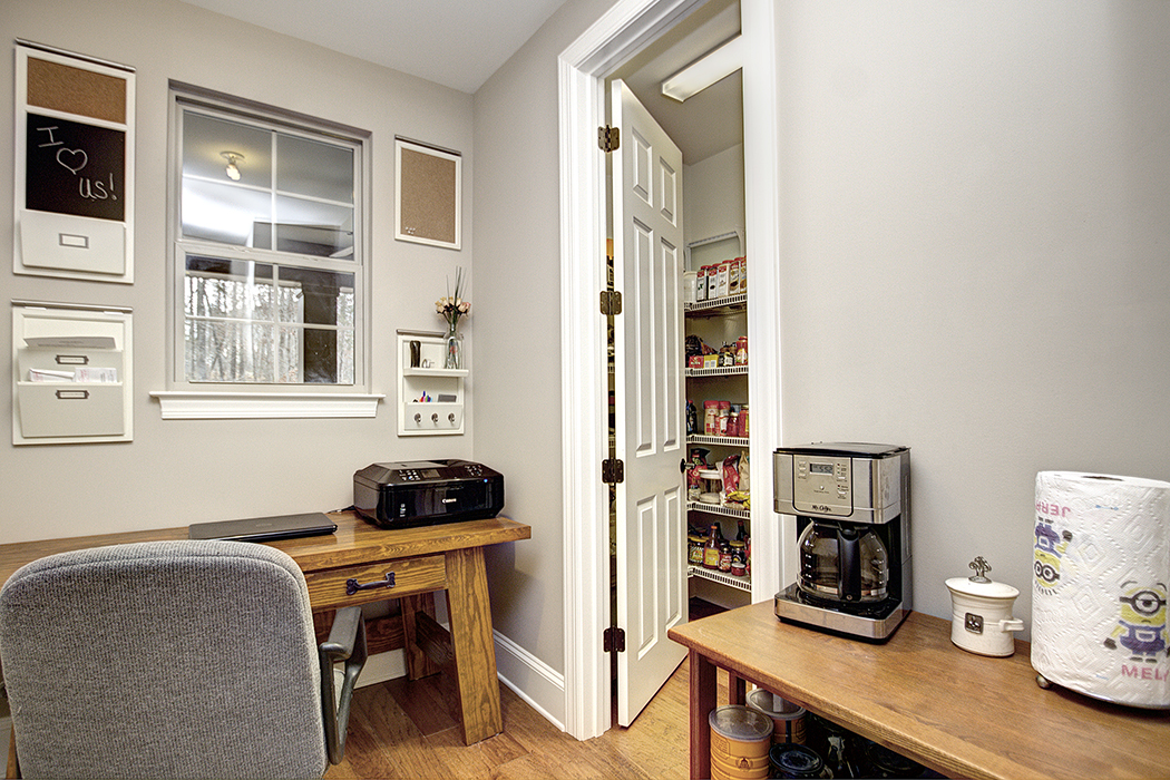Home office with gray walls light wood floors window over desk open to pantry with wire shelvinf