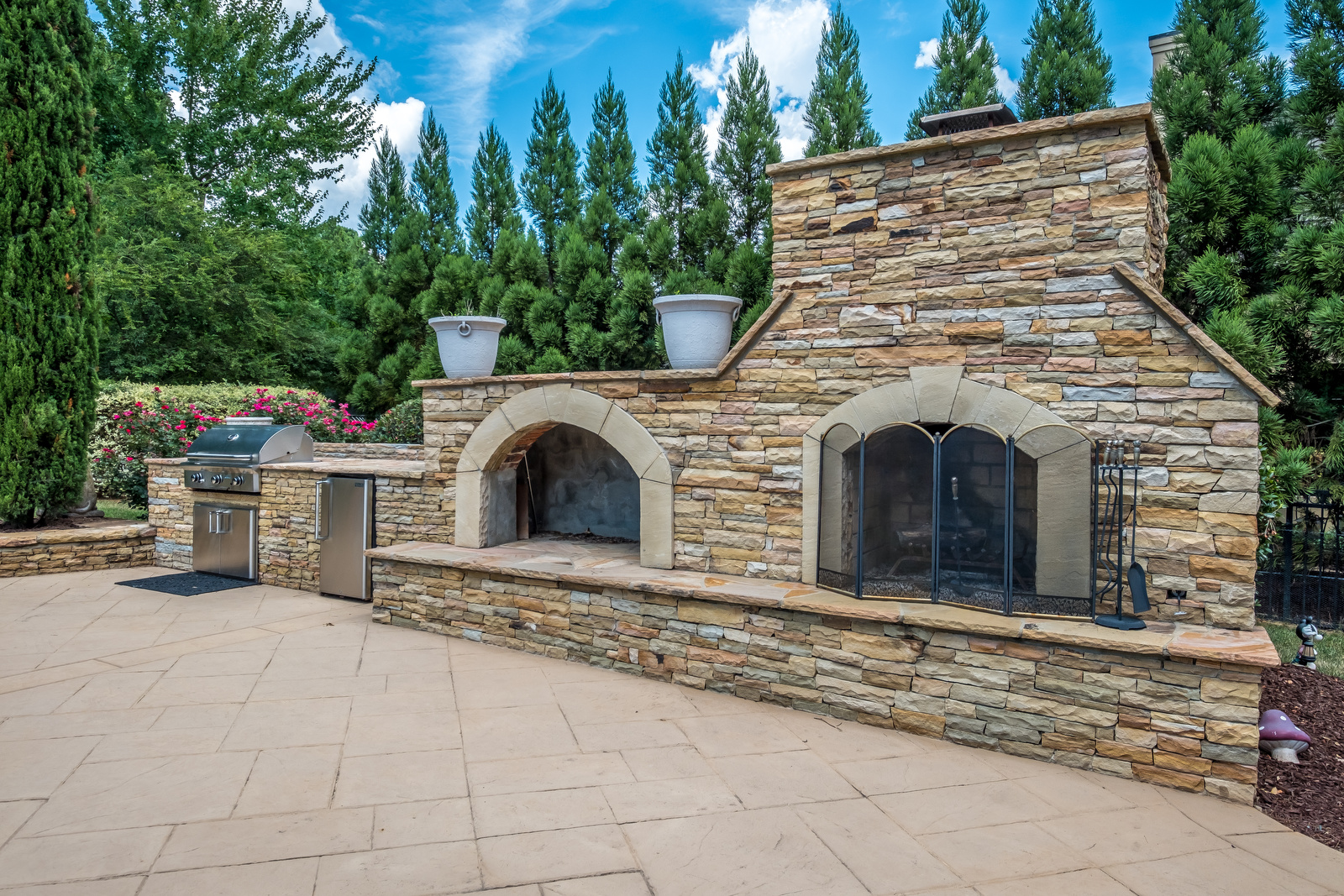 Backyard with stone grill area with built in grill beverage fridge pizza oven and fireplace with manicured trees behind it