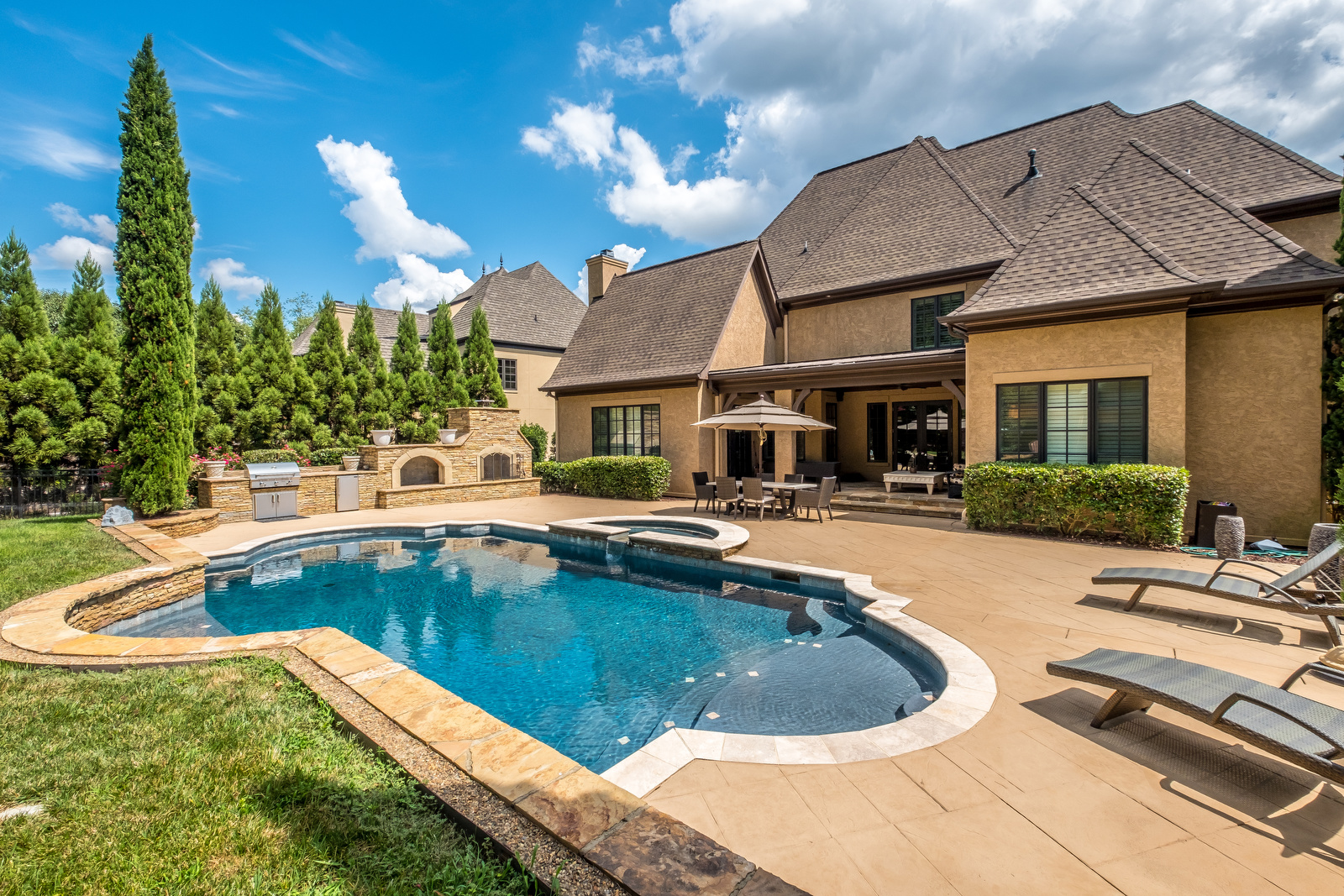 Backyard with pool with stone details eating area hot tub and built in fireplace and grill area with playground in background