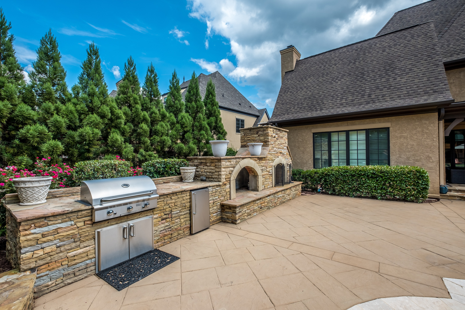 Backyard with built in grill area drink fridge pizza oven and fireplace stone details manicured trees in background