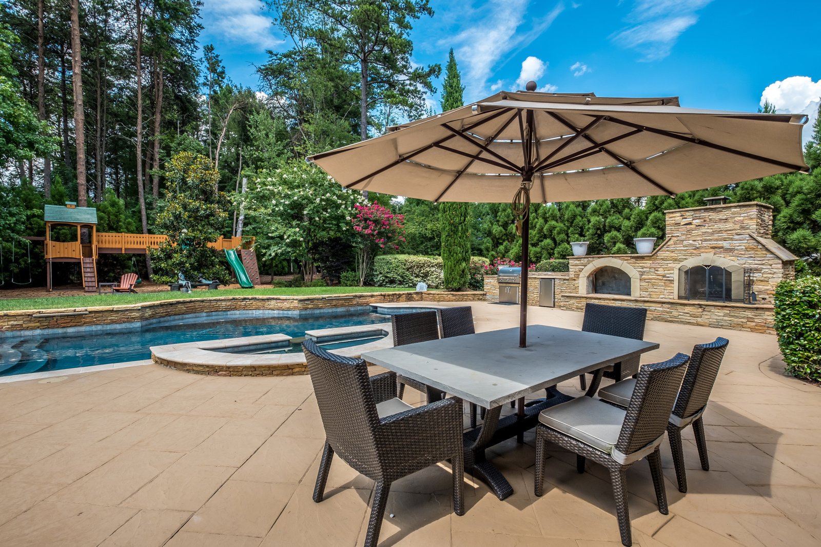 Backyard with pool with stone details eating area hot tub and built in fireplace and grill area with playground in background