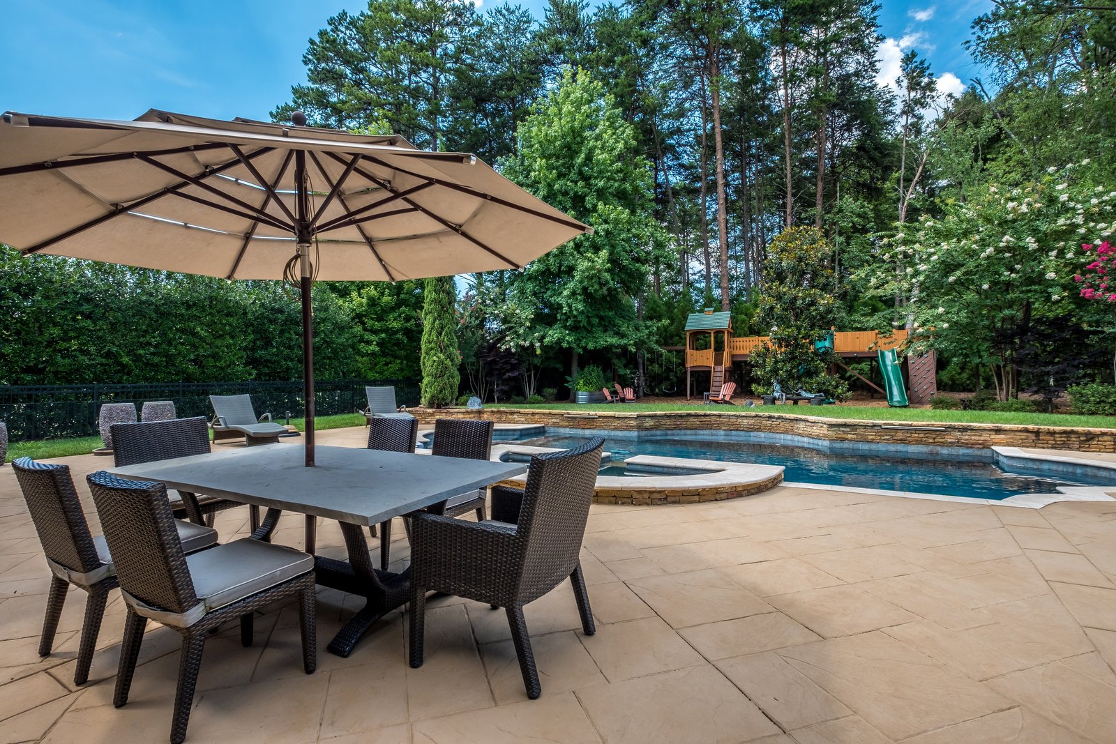 Backyard with pool with stone details eating area hot tub and built in fireplace and grill area with playground in background