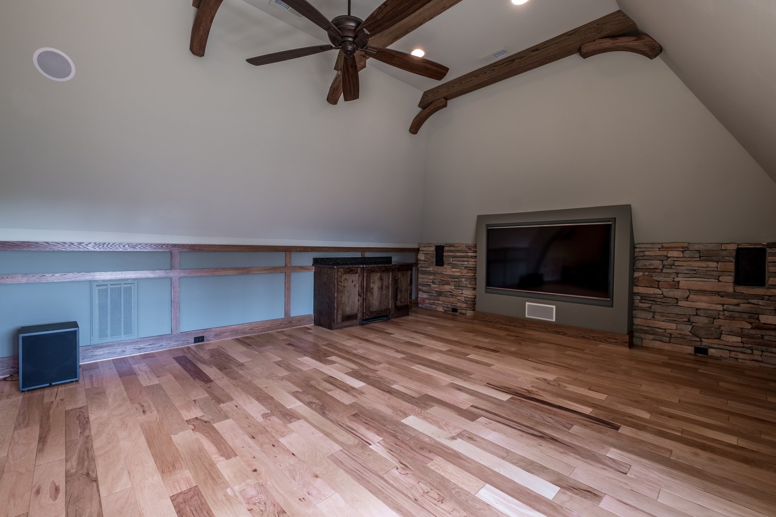 Open room with wood floors light gray walls vaulted ceilings dark wood beams dark wood fan brick wall built in TV