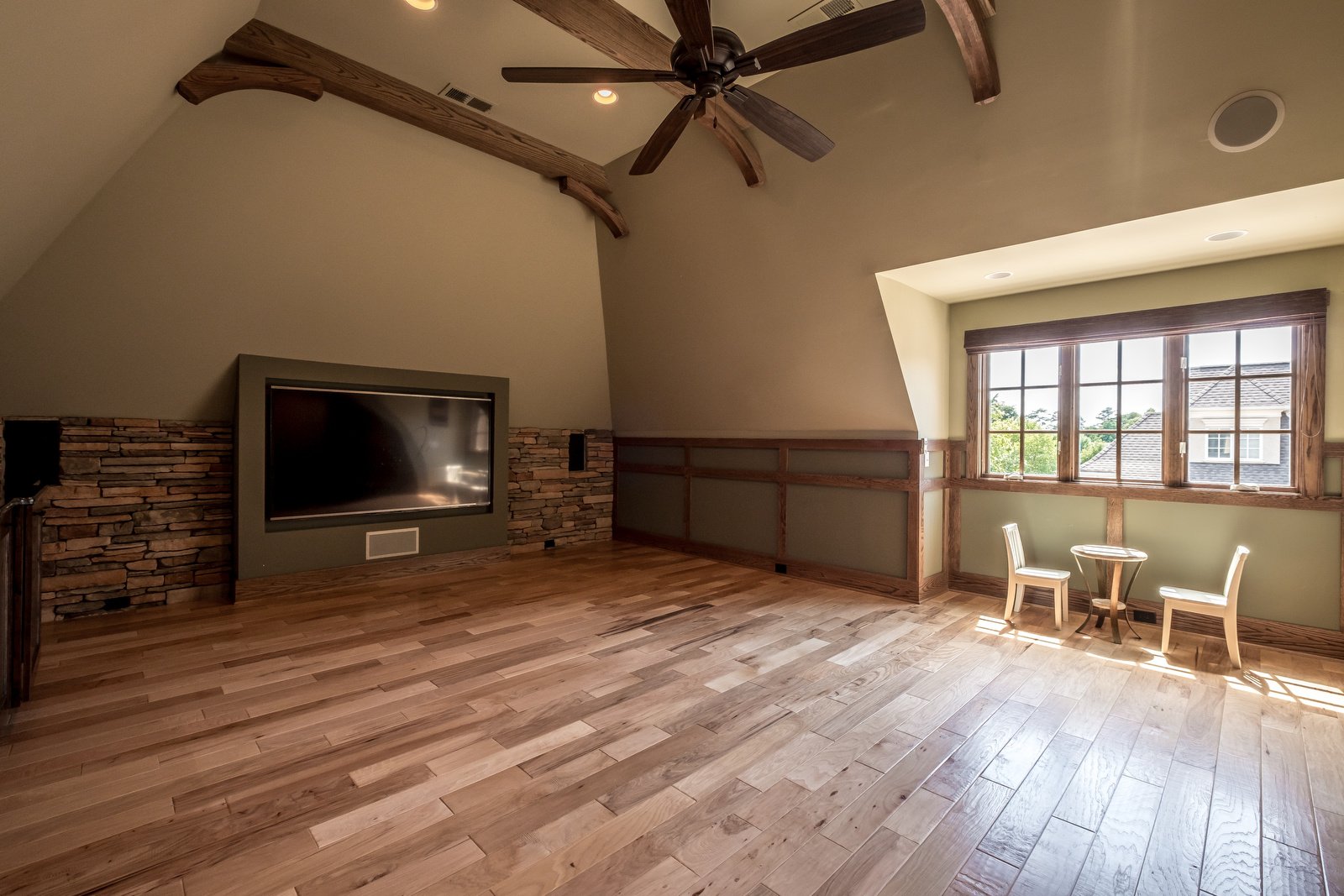 Open room with wood floors light gray walls vaulted ceilings dark wood beams dark wood fan brick wall built in TV