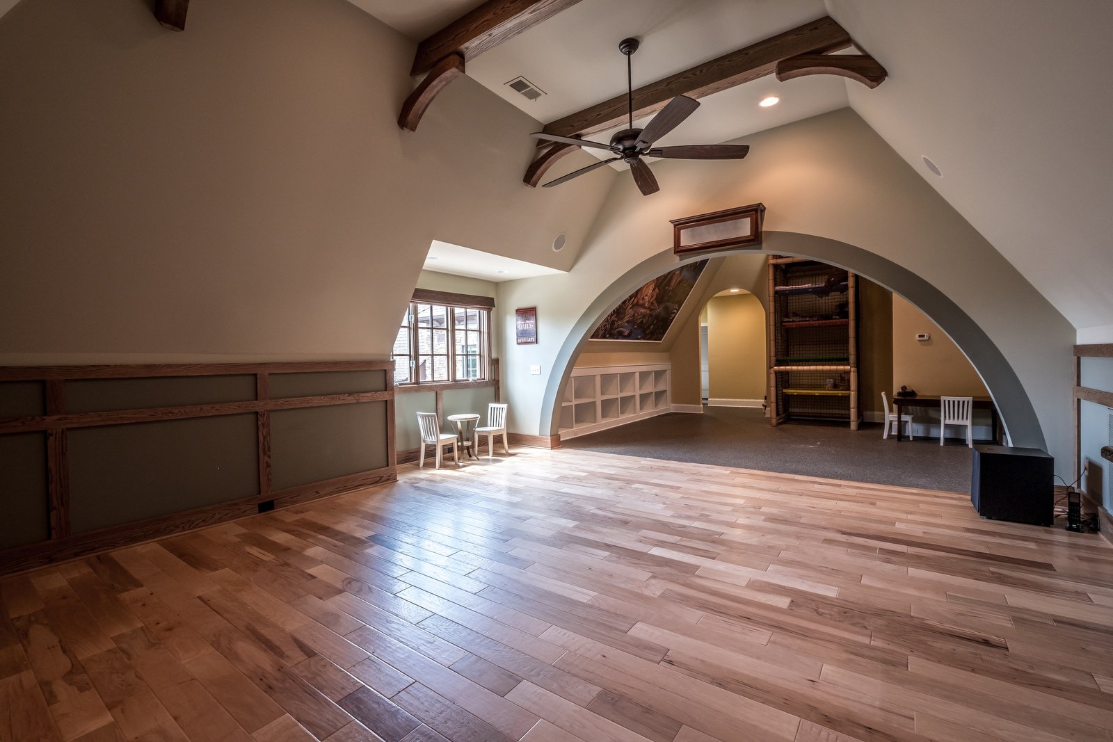 Open room with wood floors light gray walls vaulted ceilings dark wood beams dark wood fan brick wall open to playroom