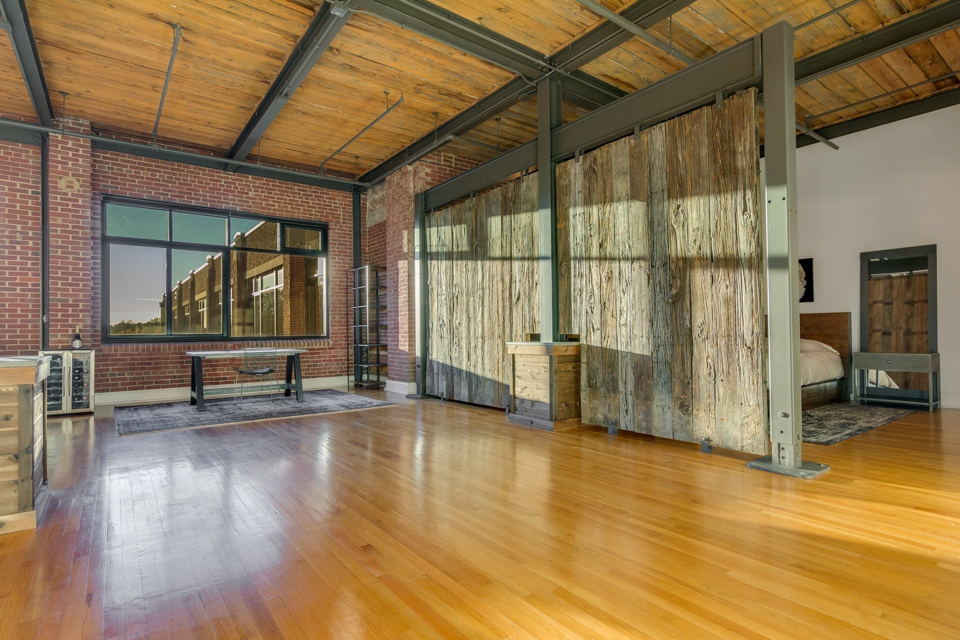 Office area open to living room with red brick wall light wood floors light wood ceiling with exposed pipes and black beams wooden partition to bedroom