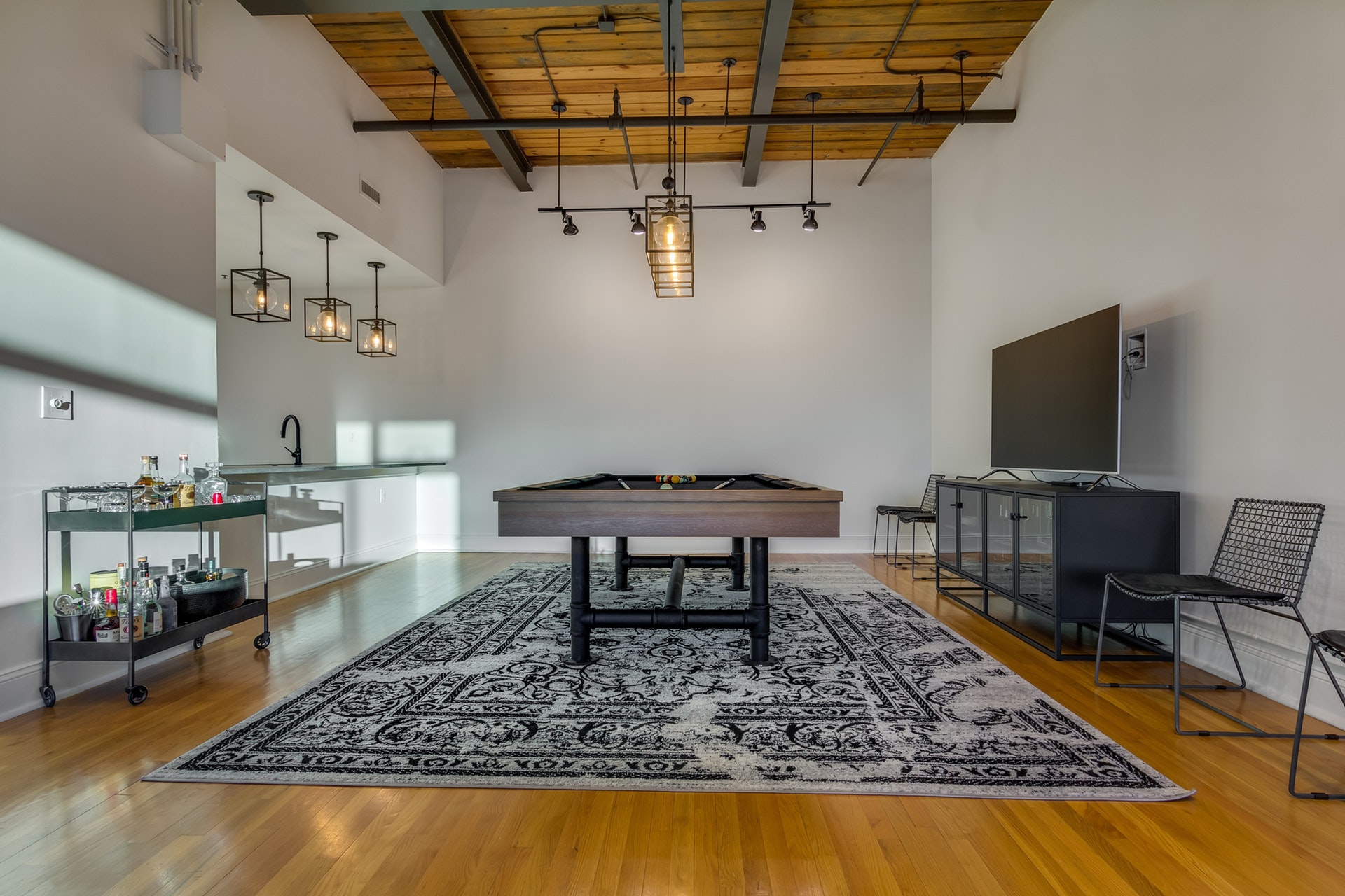 Game room with light wood floor white walls light wood ceiling exposed pipes track and pendant lighting