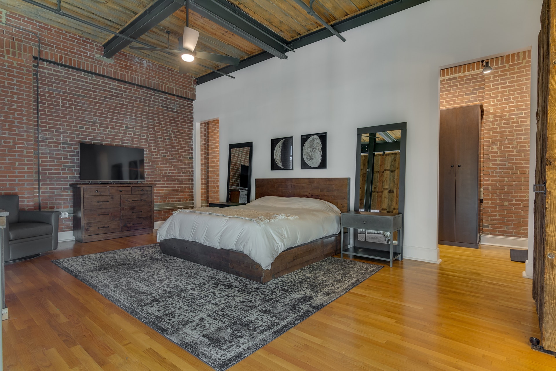 Bedroom with brick wall and white wall light wood floor open to closet wood ceiling large ceiling fan exposed pipes
