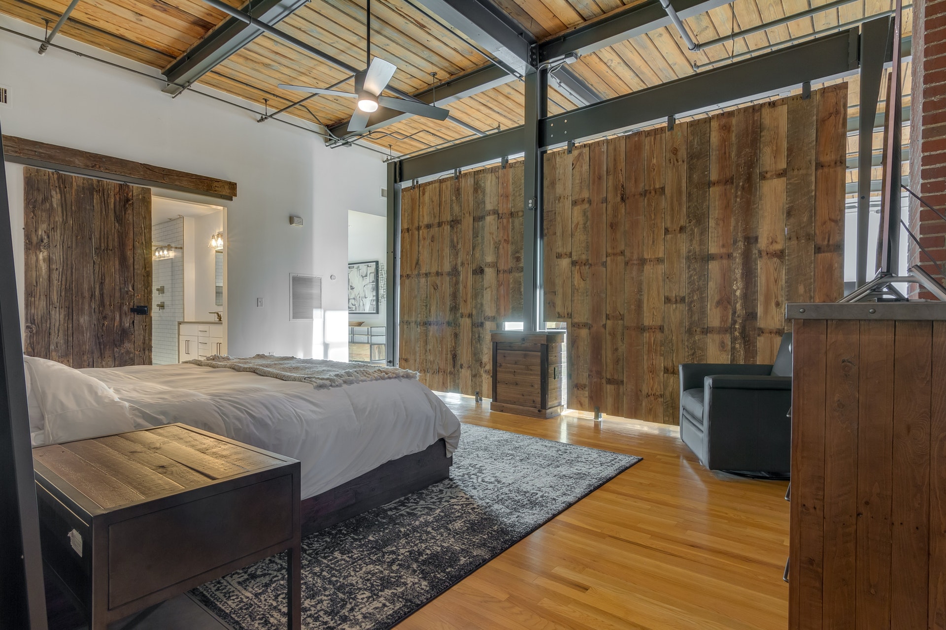 Bedroom with brick wall and white wall light wood floor wood ceiling large ceiling fan exposed pipes sliding barn door to bathroom