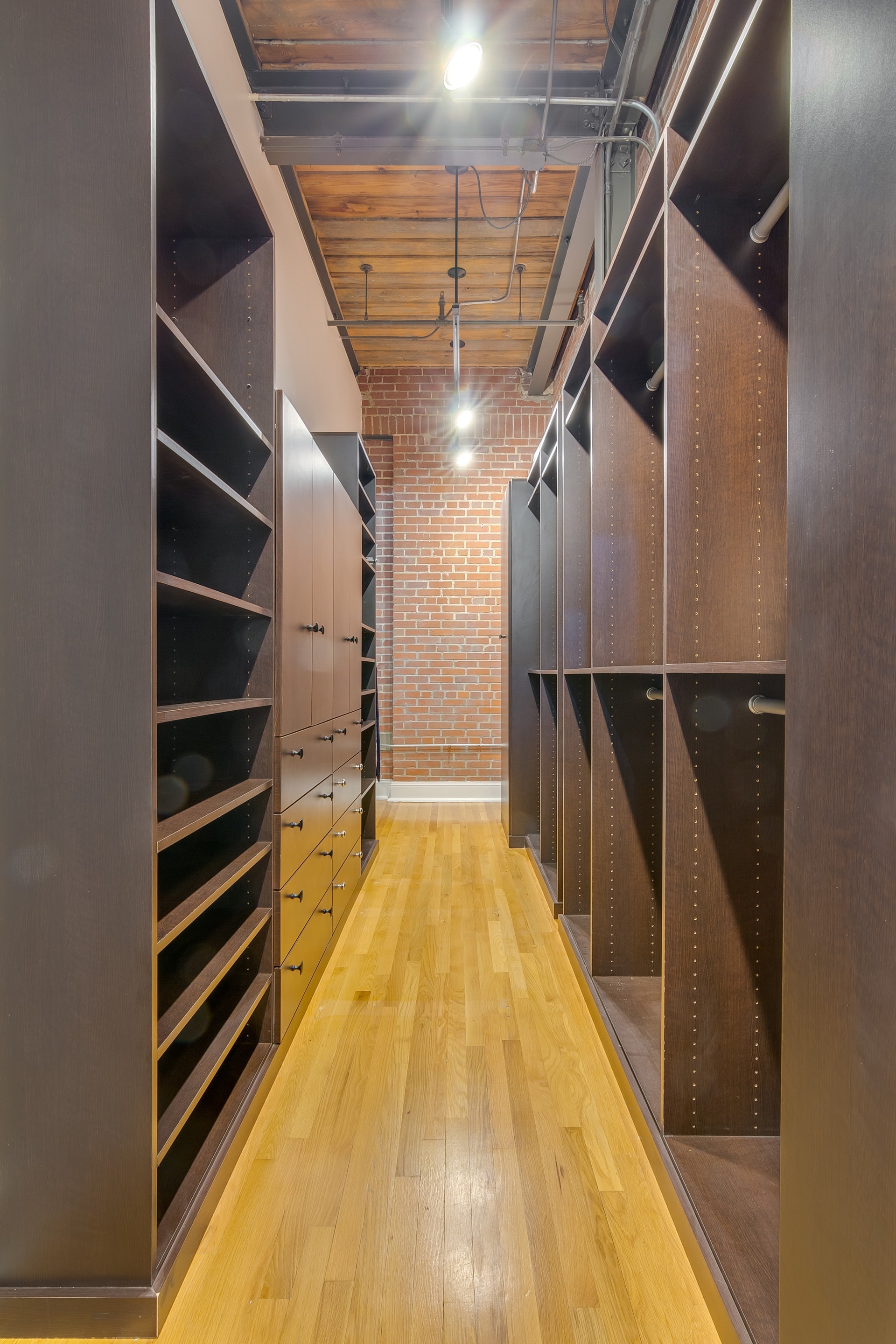 Large empty walk in closet dark brown cabinetry light wood floor exposed ceiling and red brick wall
