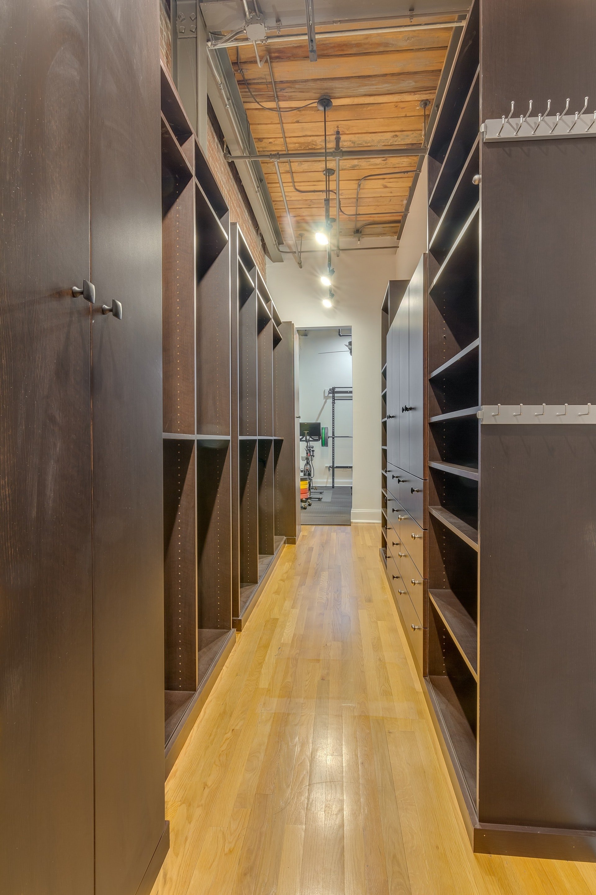 Large empty walk in closet dark brown cabinetry light wood floor open to exercise room