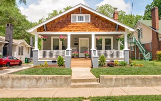 Home with gray siding and brown shingles front porch with white railing and white columns pink flowers hanging and in pots on front porch