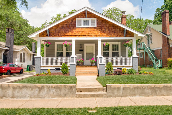 Home with gray siding and brown shingles front porch with white railing and white columns pink flowers hanging and in pots on front porch