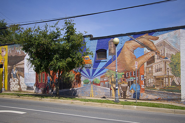Building with. mural painted on it sidewalk with tree in front and street in front of that
