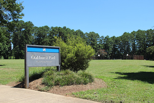 Green grass field with trees and basketball court in the background and gray and blue Oakhurst Park sign in front next to sidewalk