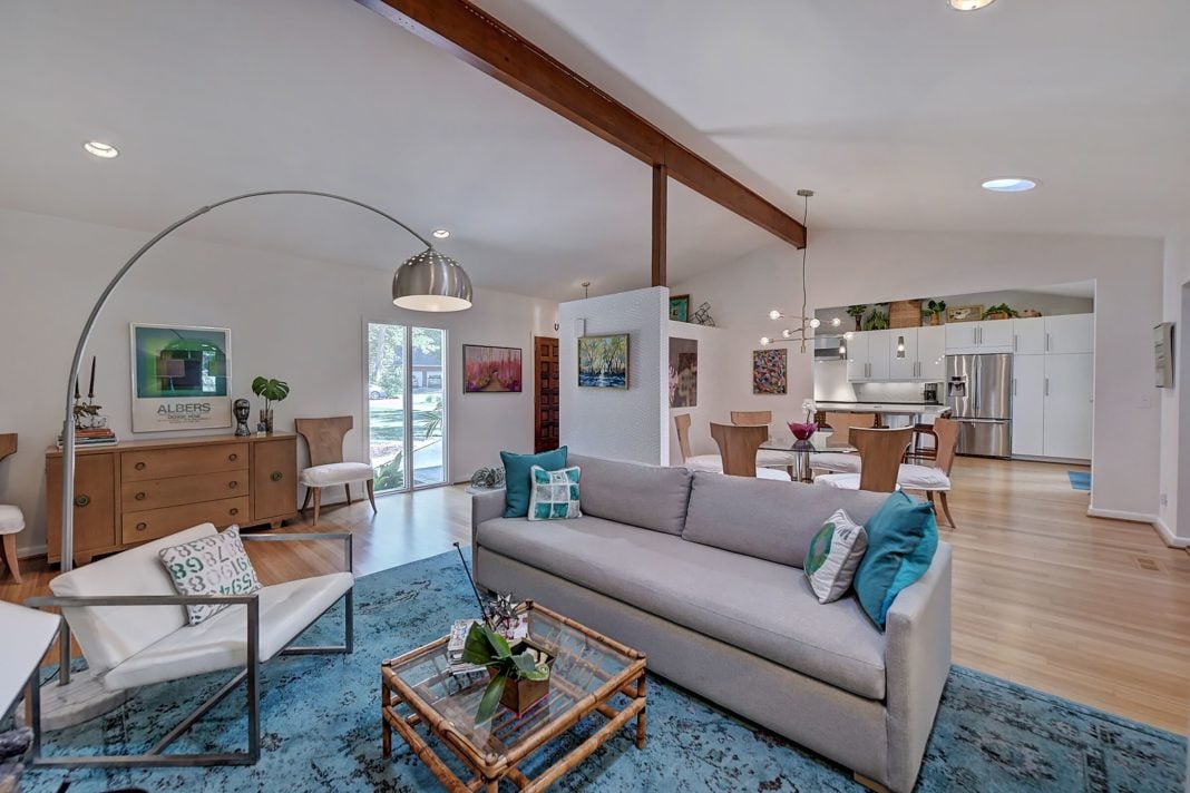 Open concept living room dining room kitchen with white walls dark wooden beam on ceiling light wood floors mid century modern furniture
