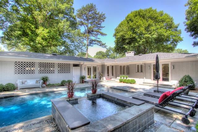Gray brick home with built in pool and hot tub with three lounge chairs and trees surrounding it