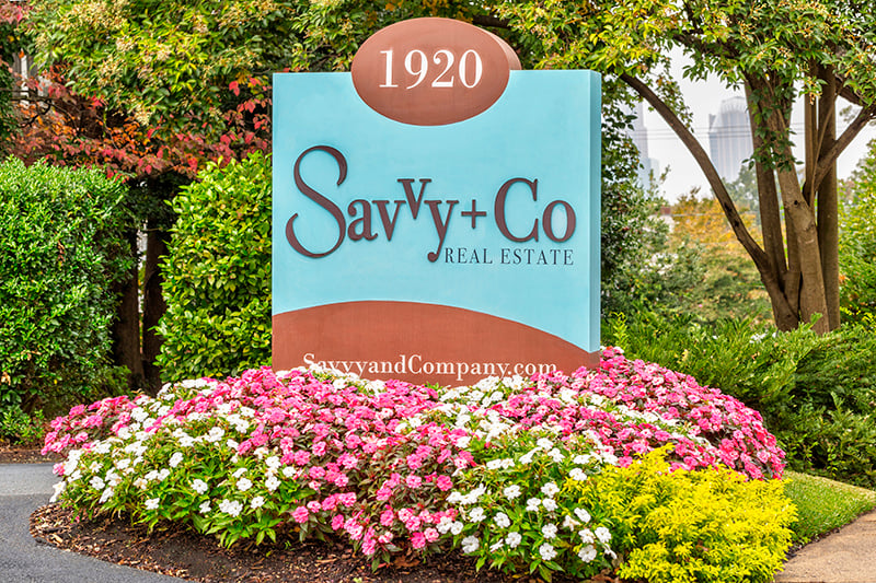 Savvy Co Real Estate blue and brown sign with pink and yellow flowers planted around it