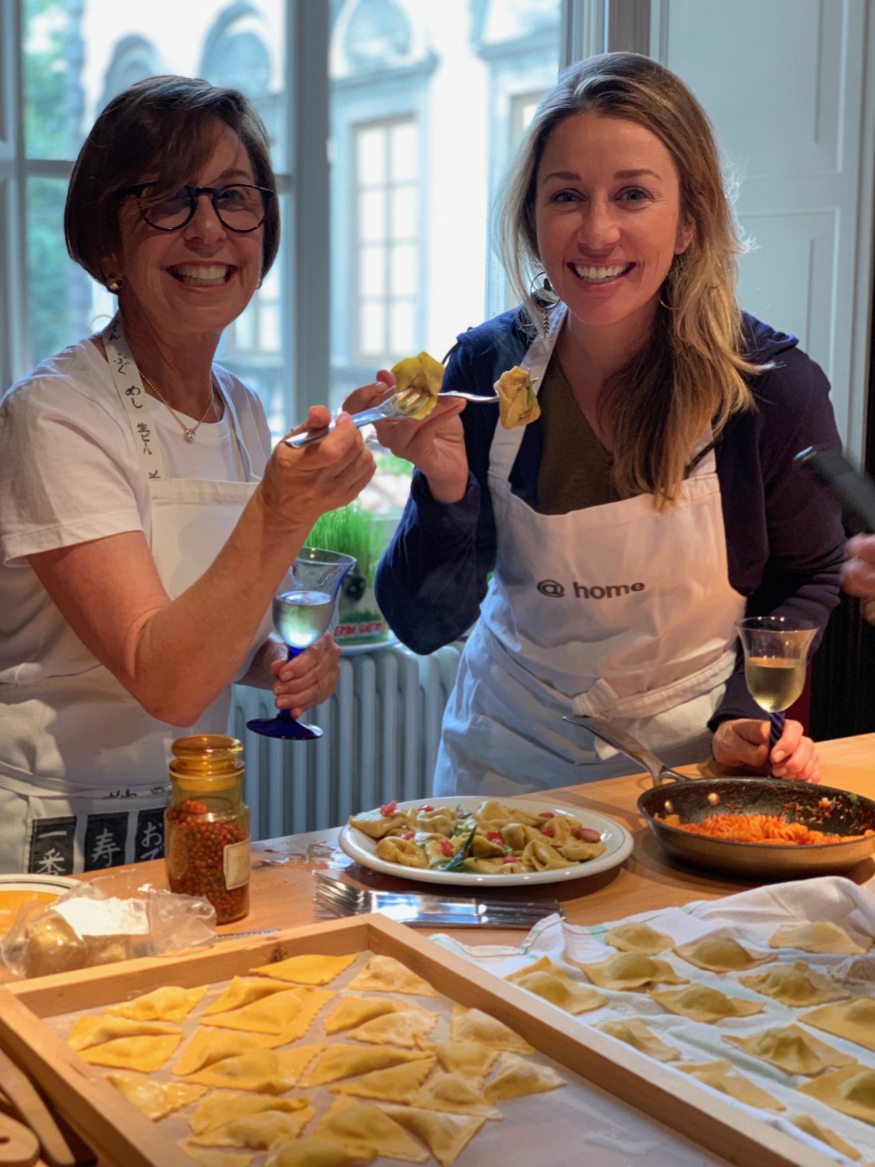 Sarah Martin and Client in Italy_1 Sarah Martin wearing a white apron standing with a lady wearing a white apron with homemade pasta around them both holding forks with pasta on them