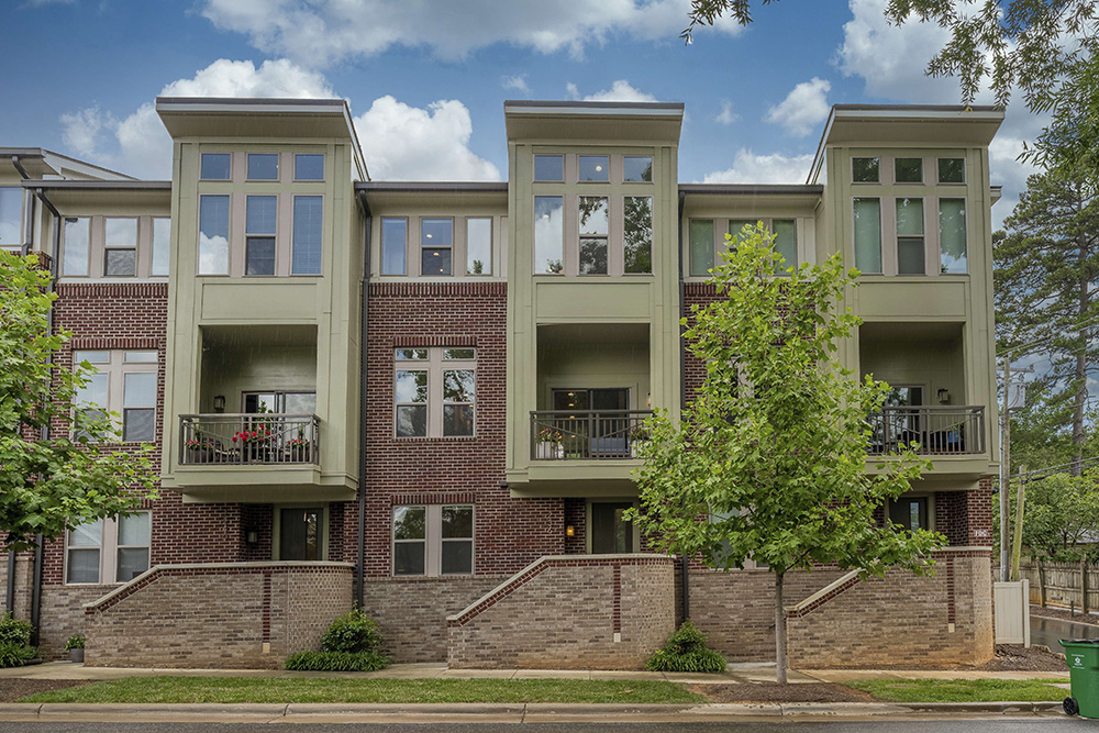Three story condo building with green exterior and red and brown brick with porches and high windows and sidewalk in front with trees lining it