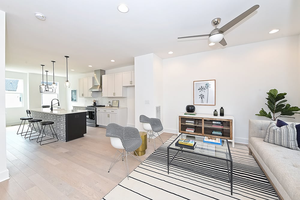 Living room with white walls and light wood floors recessed lighting and silver ceiling fan open to kitchen with white cabinets and gray tiled island