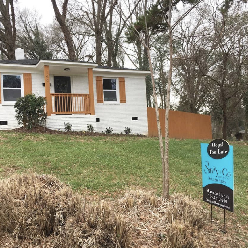 White painted brick home with brown shutters and columns and railings with fenced in backyard and front yard with green grass and a Savvy Co Real Estate sign