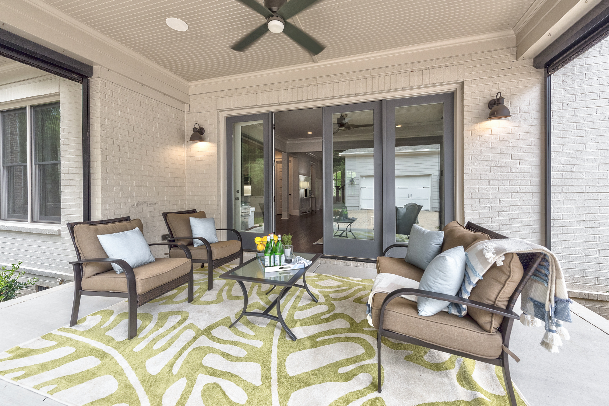 Back porch with white painted bricks and gray framed windows and glass doors with concrete floor and brown patio furniture with white and green rug