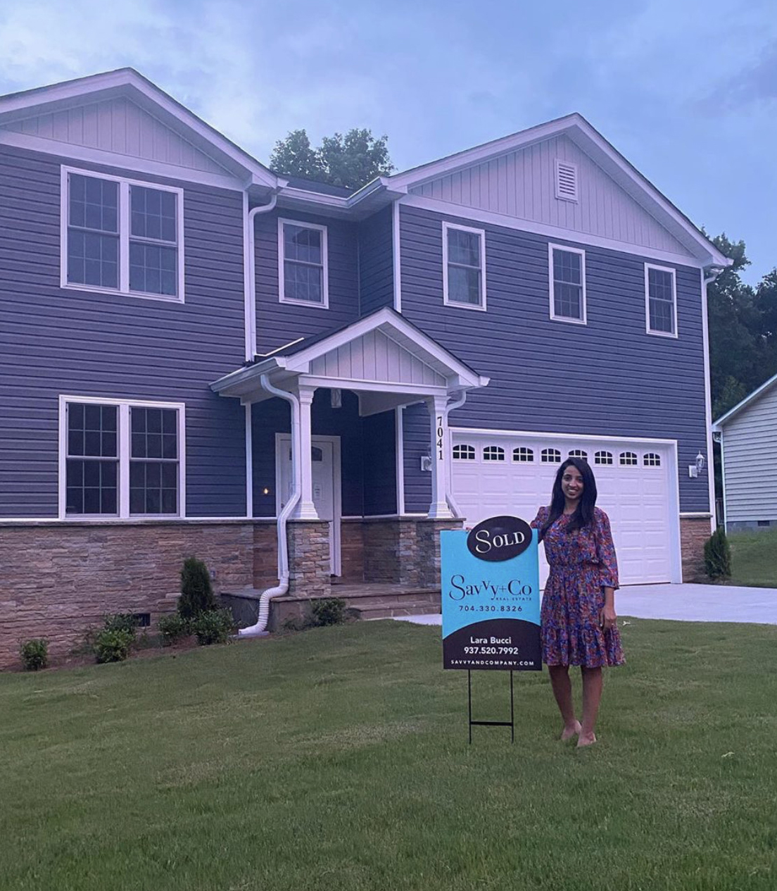 Lara Bucci wearing a long sleeve red and blue floral dress standing in the front yard of a two story home with a blue and brown Savvy sold sign