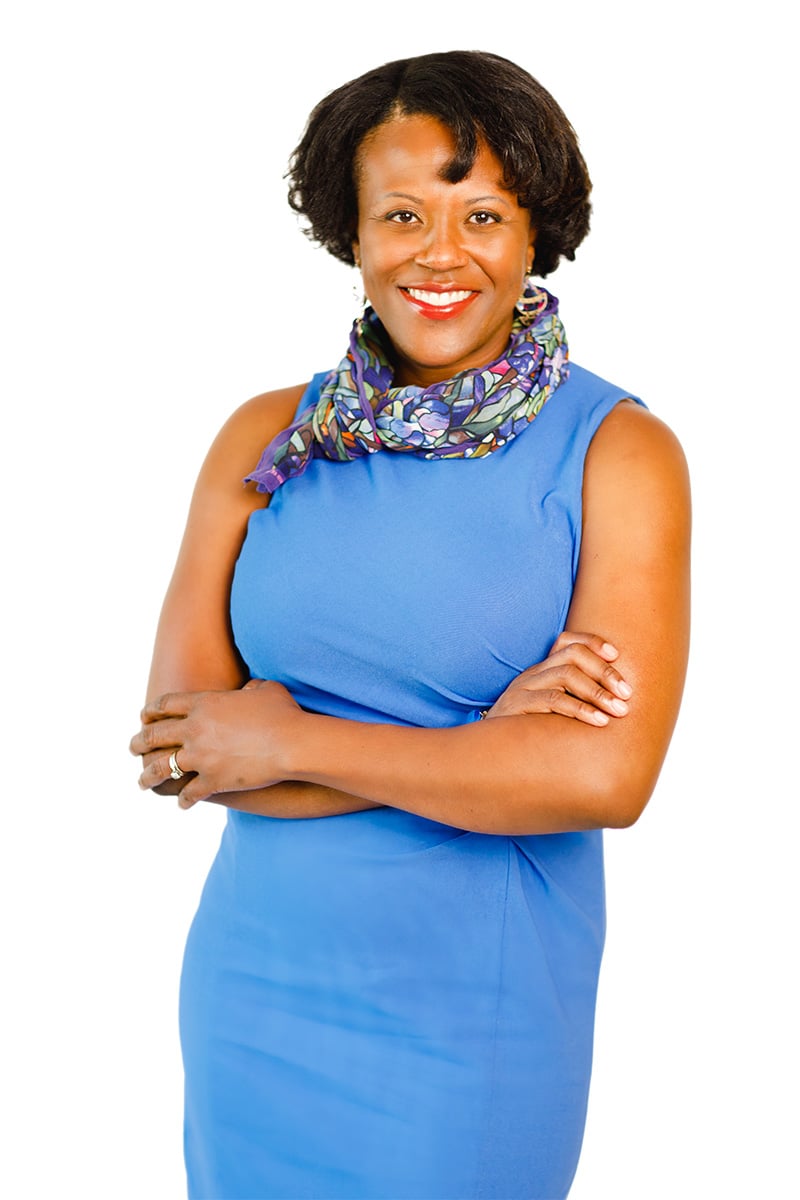Kim Parati wearing a blue sleeveless dress and blue floral scarf standing in front of a white backdrop