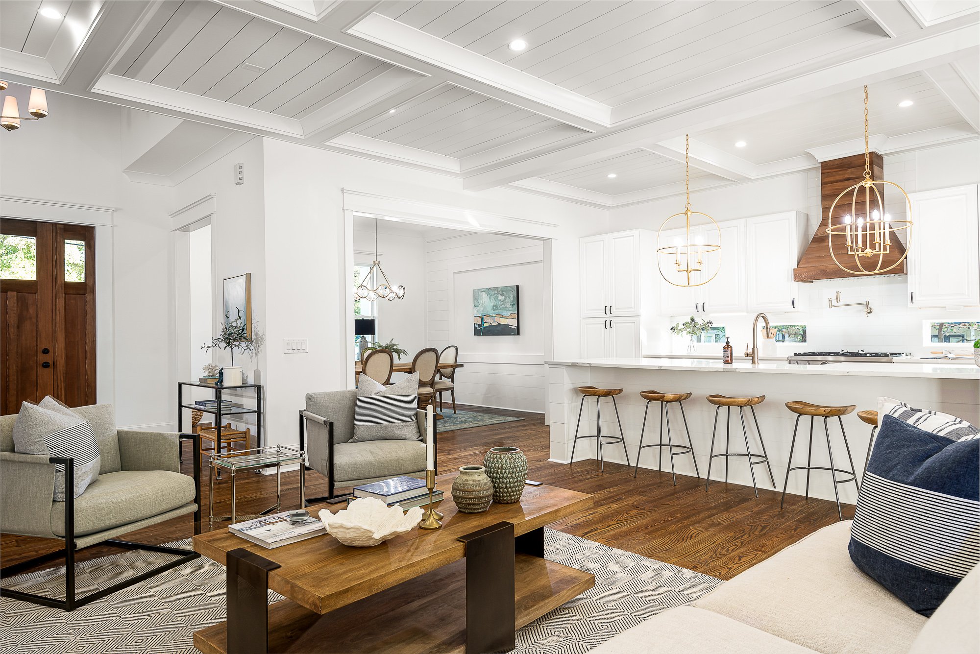 Living room with white walls and medium wood floors and wood front door open to kitchen with white cabinets stainless steel appliances and golf light fixtures